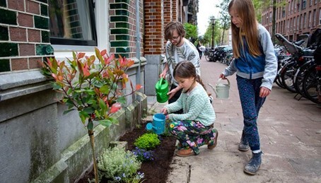 Kinderen in een stadstraat een de rand van een buitengevel een geveltuin. Ze geven water aan de geveltuin.