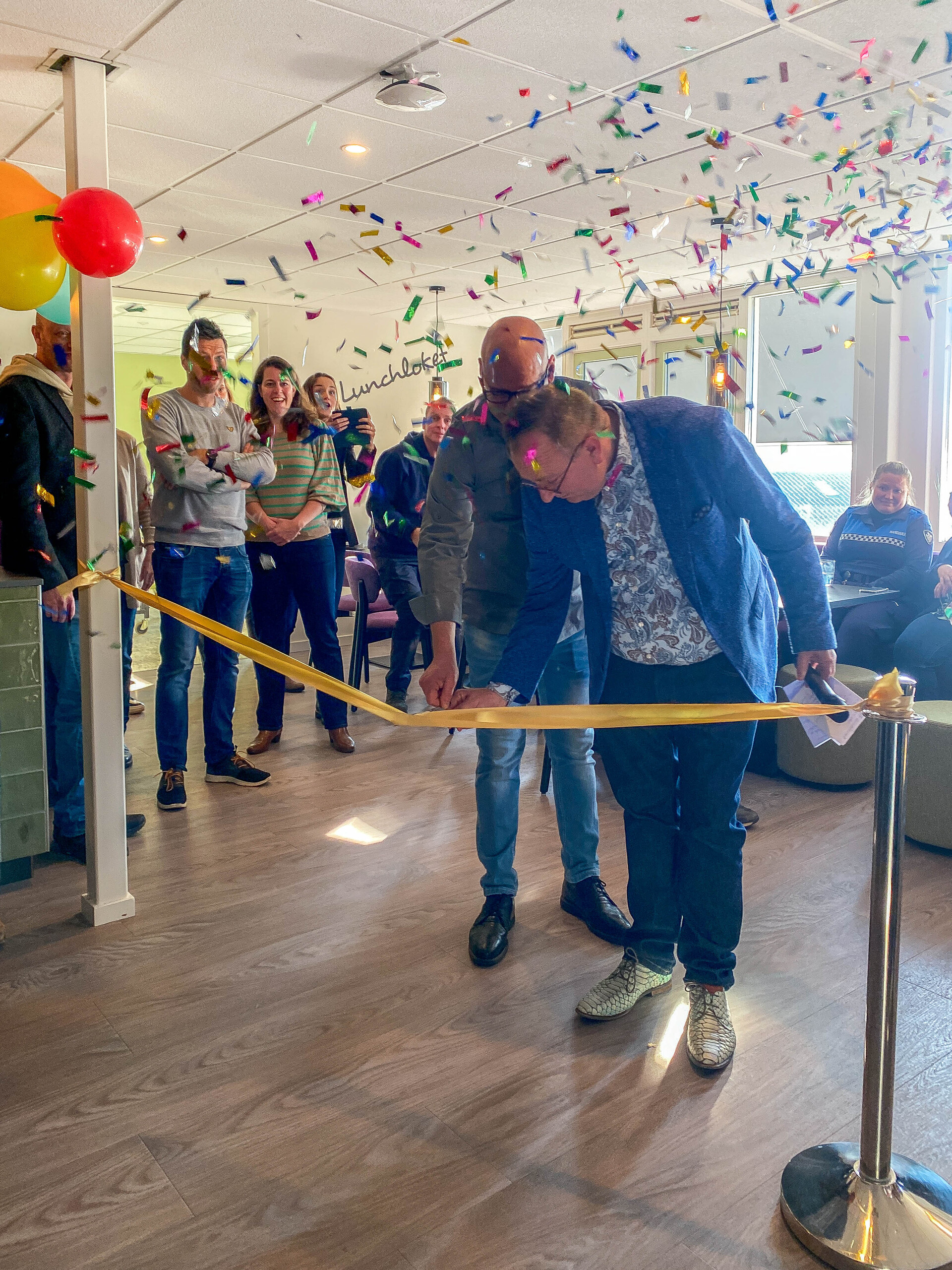 Foto van de opening van de kantine tijdens het doorknippen van het lint. Gemeentesecretaris knipt het lint door, samen met Sjoerd, een van de medewerkers van Rijnvicus.