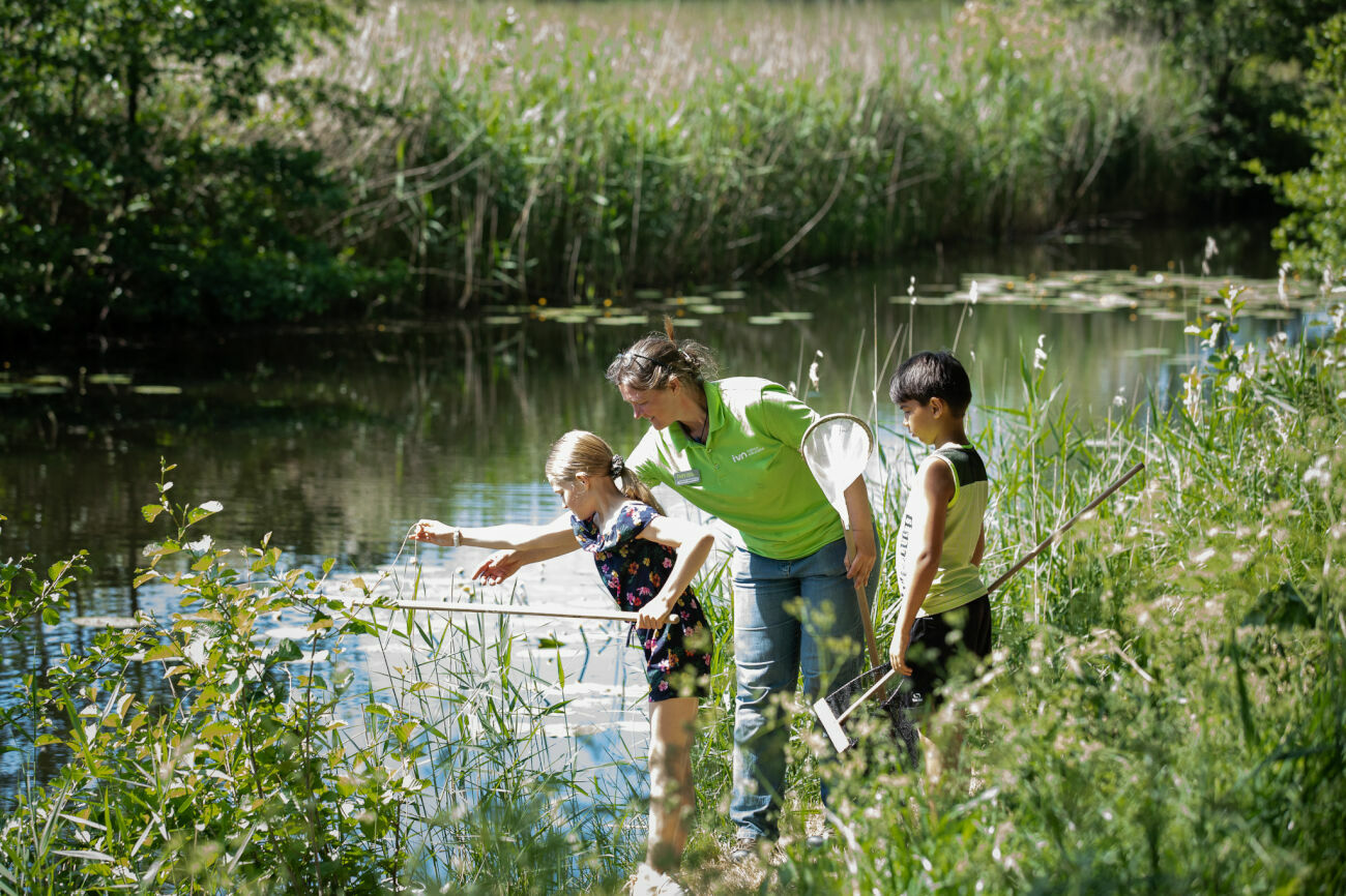 foto van twee kinderen samen met een medewerker van IVN die aan de slootkant staan. De medewerker legt verschillende dingen uit over de waterkant en de bewoners daarvan.