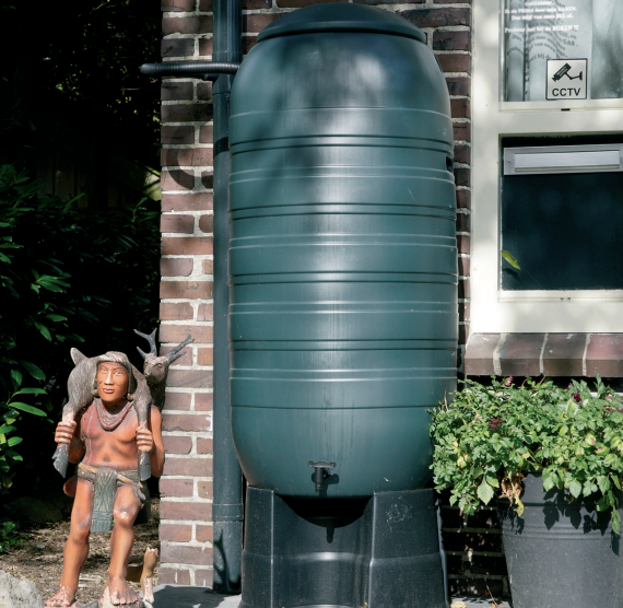 Een groene regenton geplaatst naast een bakstenen gebouw met een klein inheems thema standbeeld en potplanten eromheen.